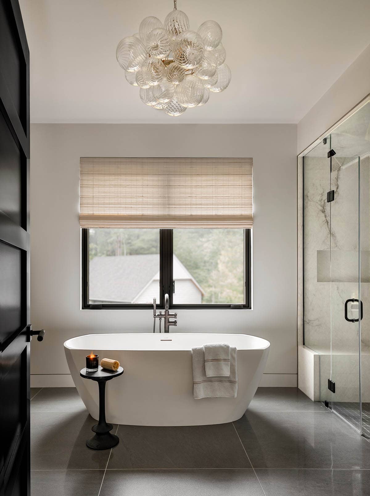 Freestanding tub under a bubble glass chandelier by a window, beside a glass shower