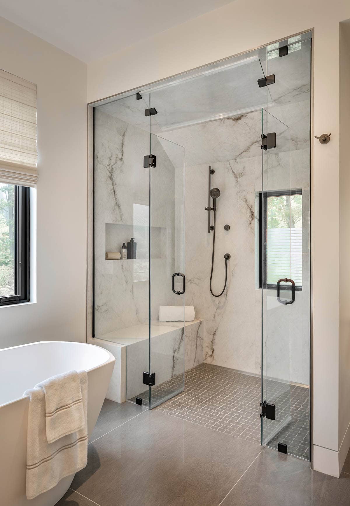 Steam shower with white marble walls, black fixtures, a bench, and a window, next to the tub