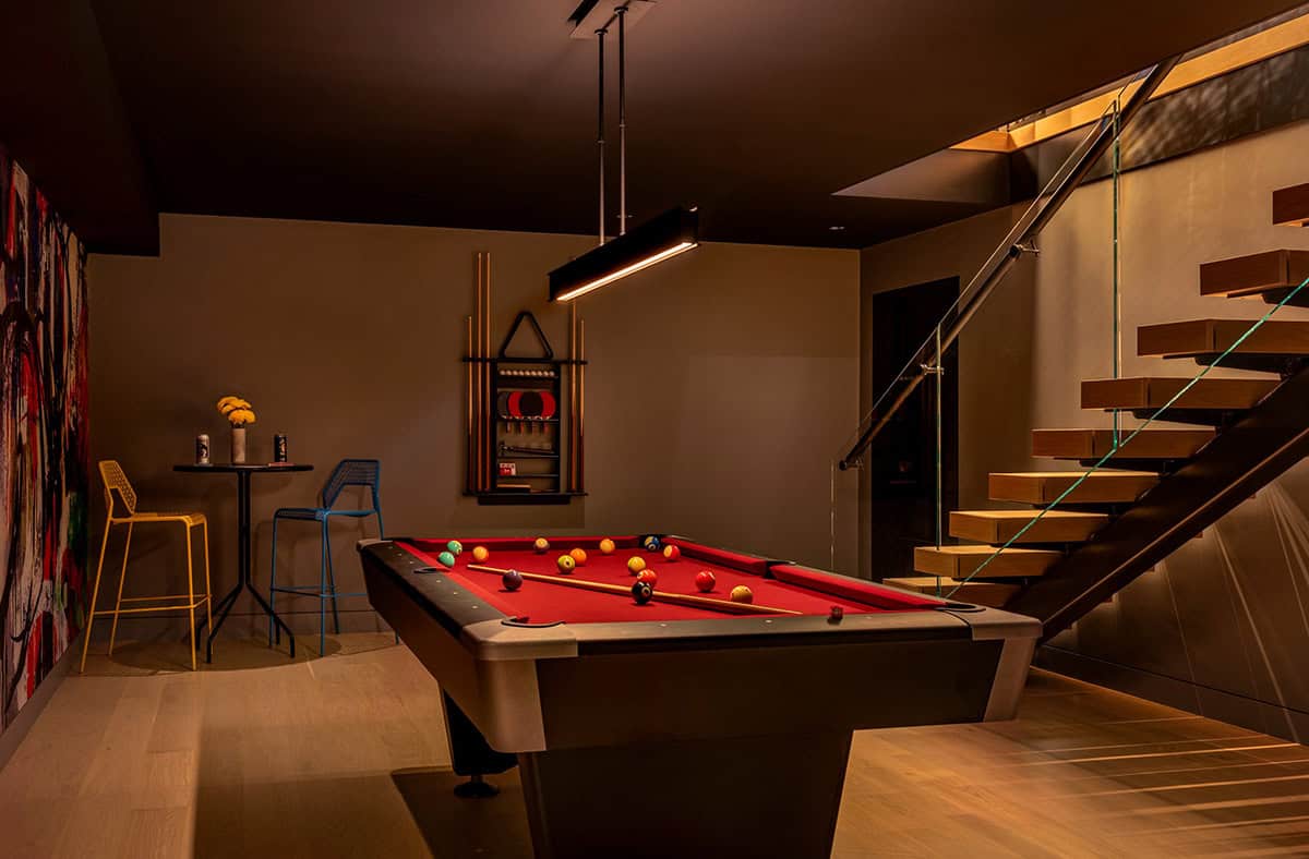 Red pool table under a linear light, with a cue rack, bar table with stools, and the floating stairs
