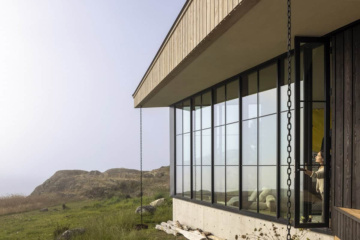 Corner of the house with a steel-framed window wall, a deep roof overhang, and a rain chain hanging over a grassy bluff