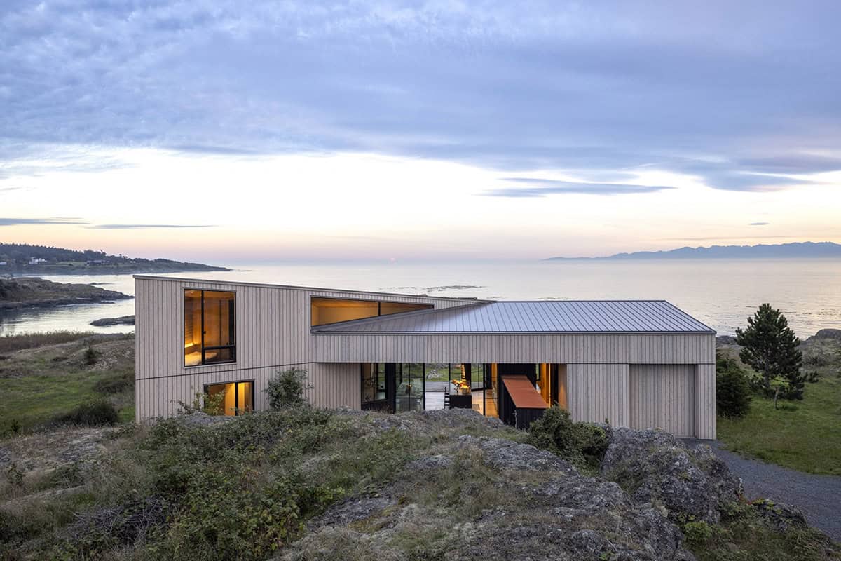 Wood-clad house on a rocky headland at dusk, with warm interior light and a view of the water and distant mountains