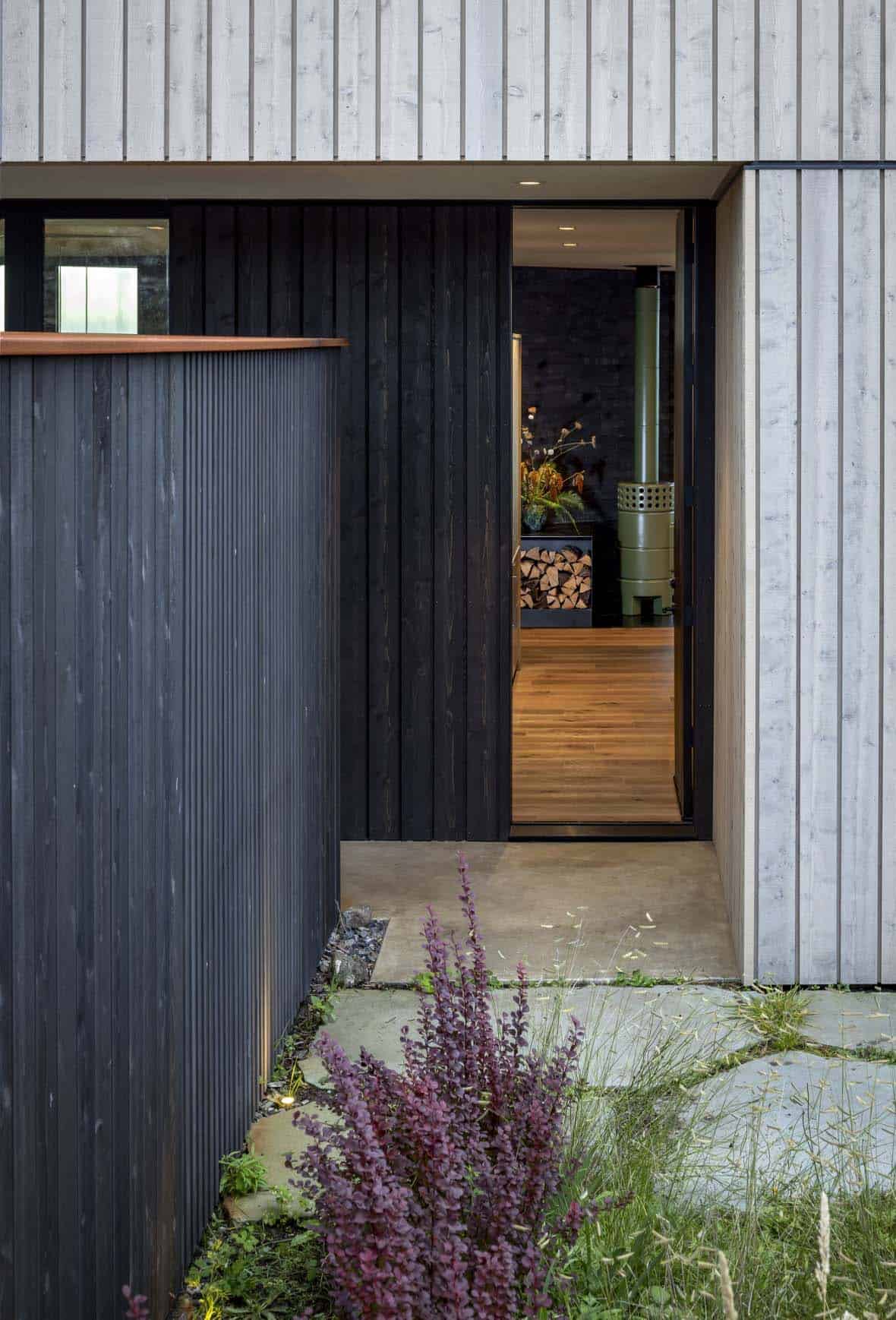 Entry nook between charred black siding and pale wood siding, with a view through the open door to a green wood stove