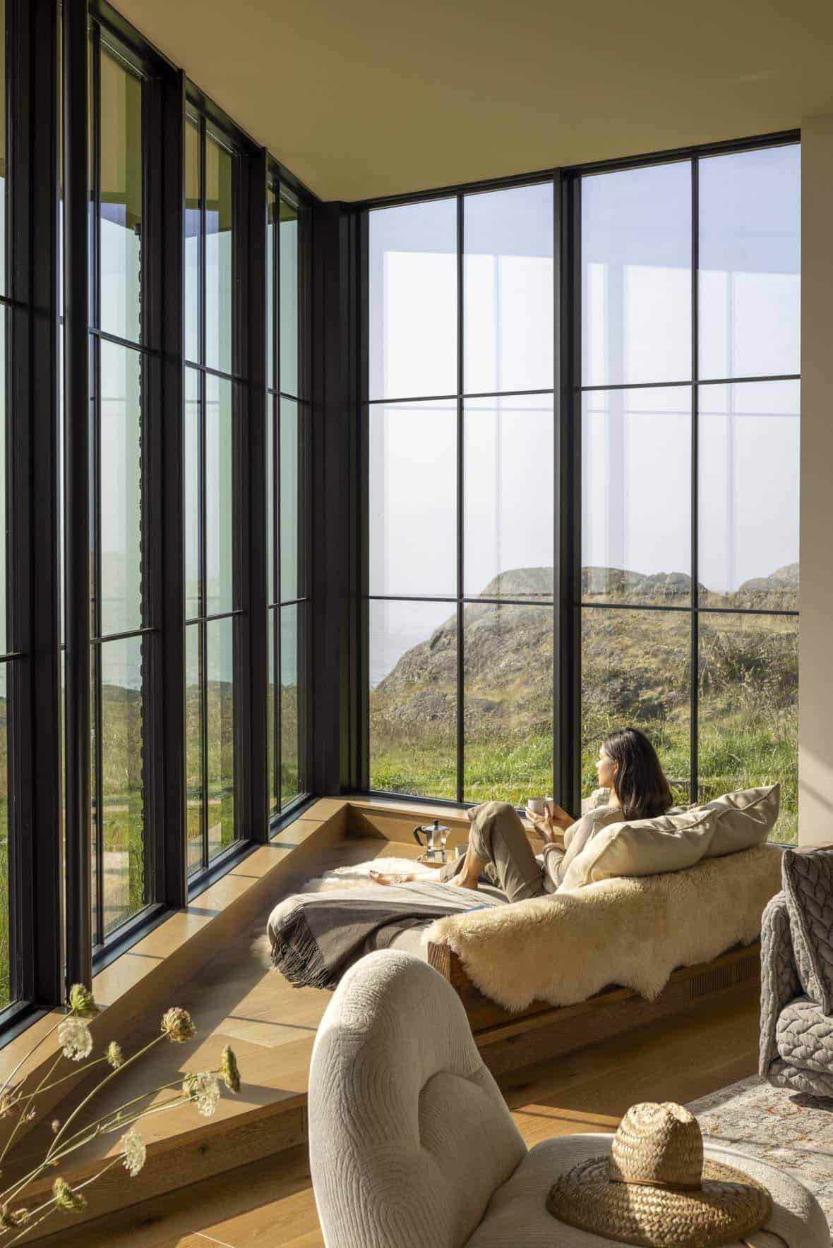Woman with a coffee cup relaxing on a sheepskin-covered daybed in a sunken window corner overlooking the coastline
