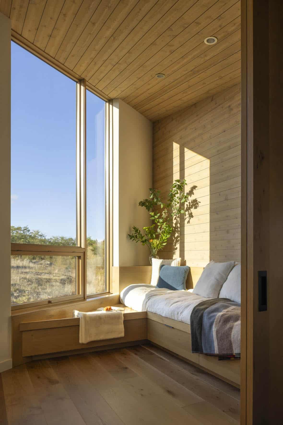 Sunlit window seat nook with built-in storage, light wood paneling, and a vase of greenery