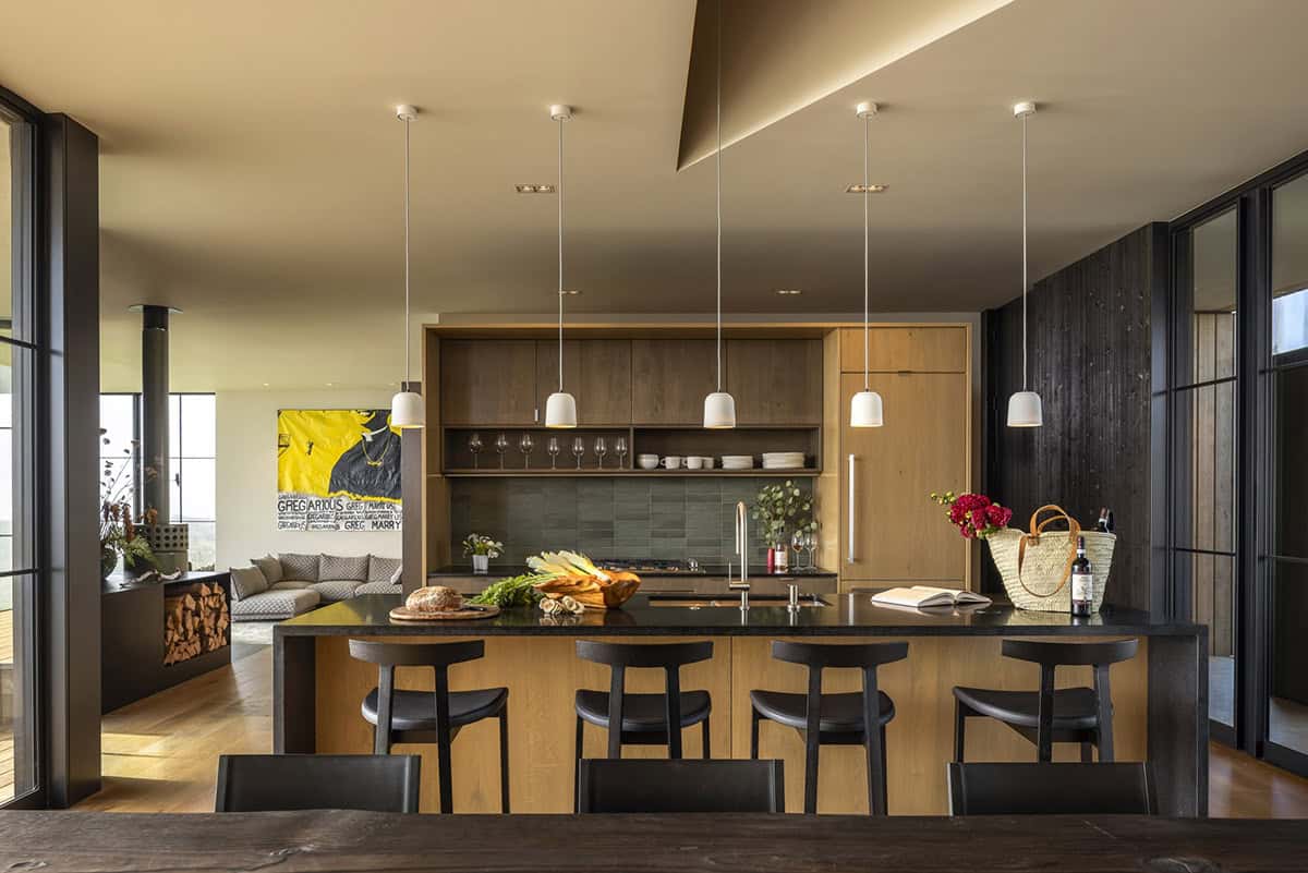 Open kitchen with a dark-topped oak island, black stools, pendant lights, and a yellow painting in the background