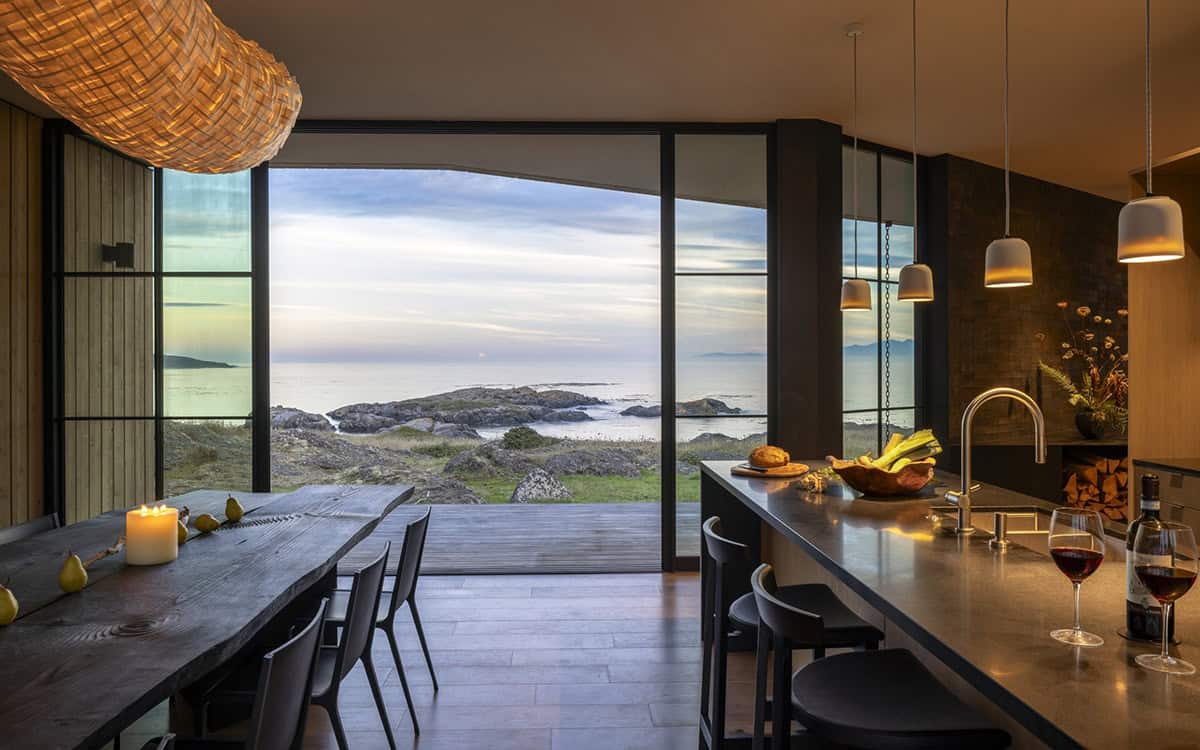 Dining table and kitchen island facing a wall of glass with a deck and sunset view of the sea