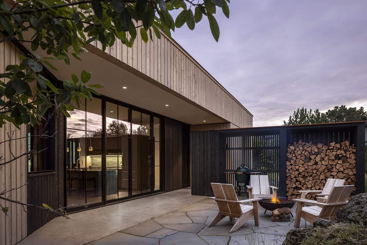 Stone patio at dusk with a fire pit, Adirondack chairs, a grill, and stacked firewood beside the glass doors
