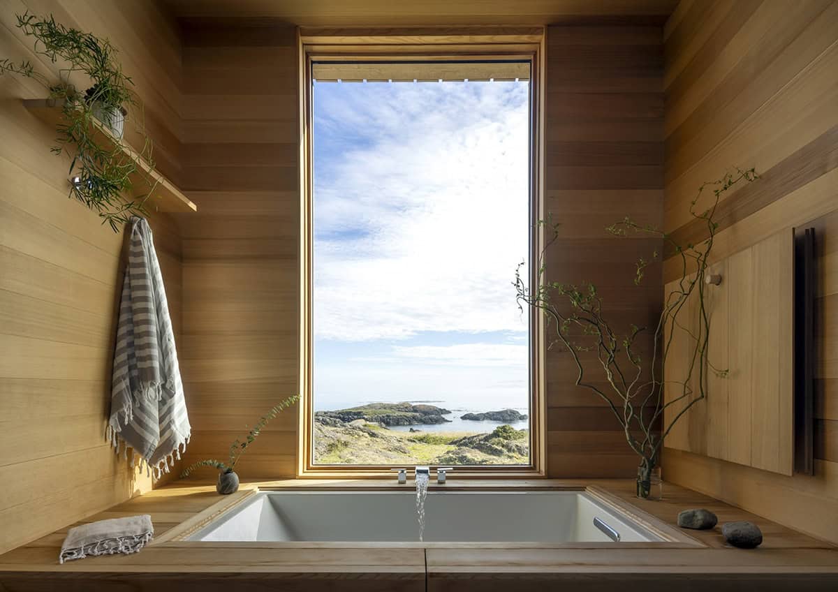 Built-in wooden soaking tub beneath a tall window framing the rocky shoreline