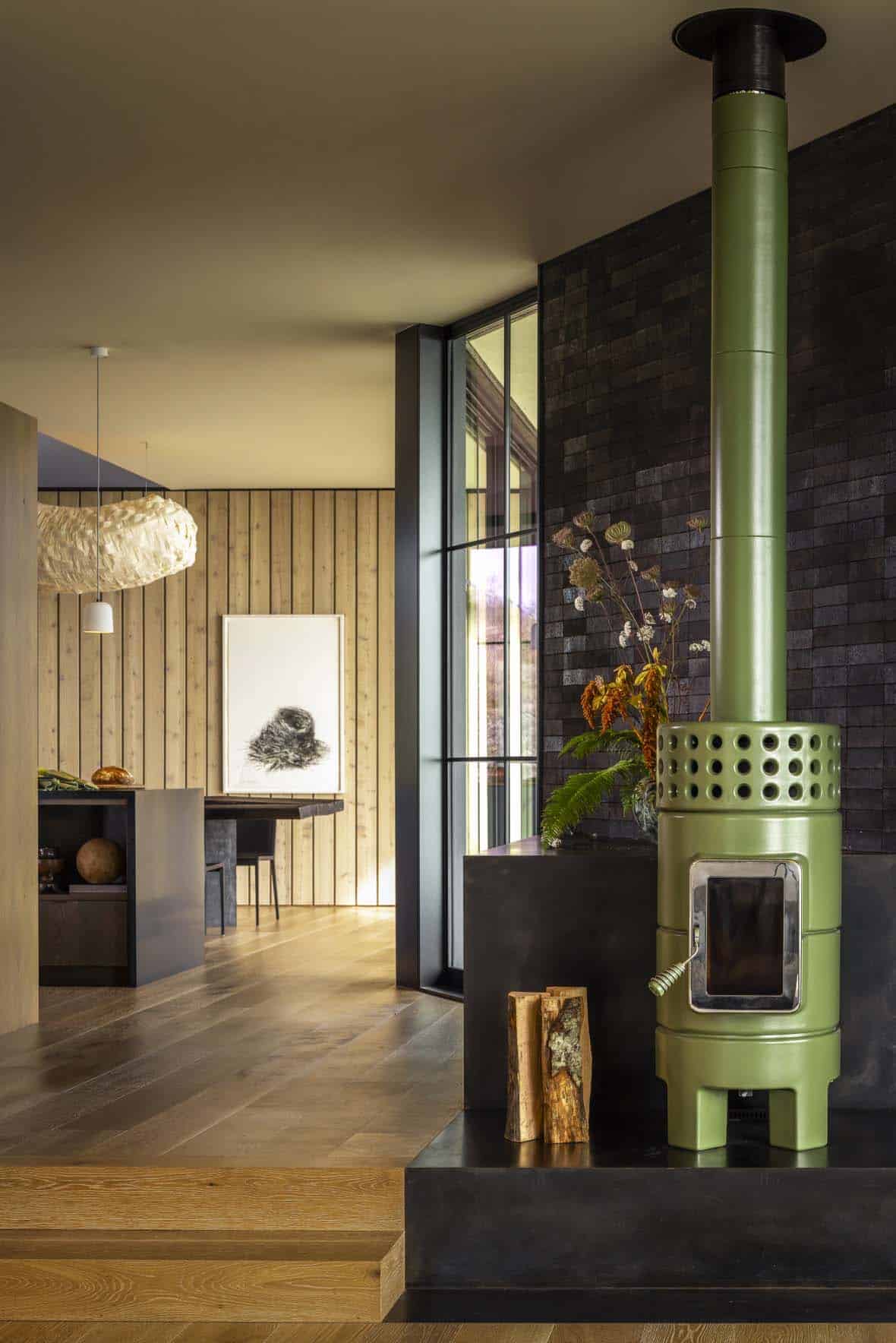 Green ceramic wood stove against a dark tiled wall, with the kitchen and dining area beyond