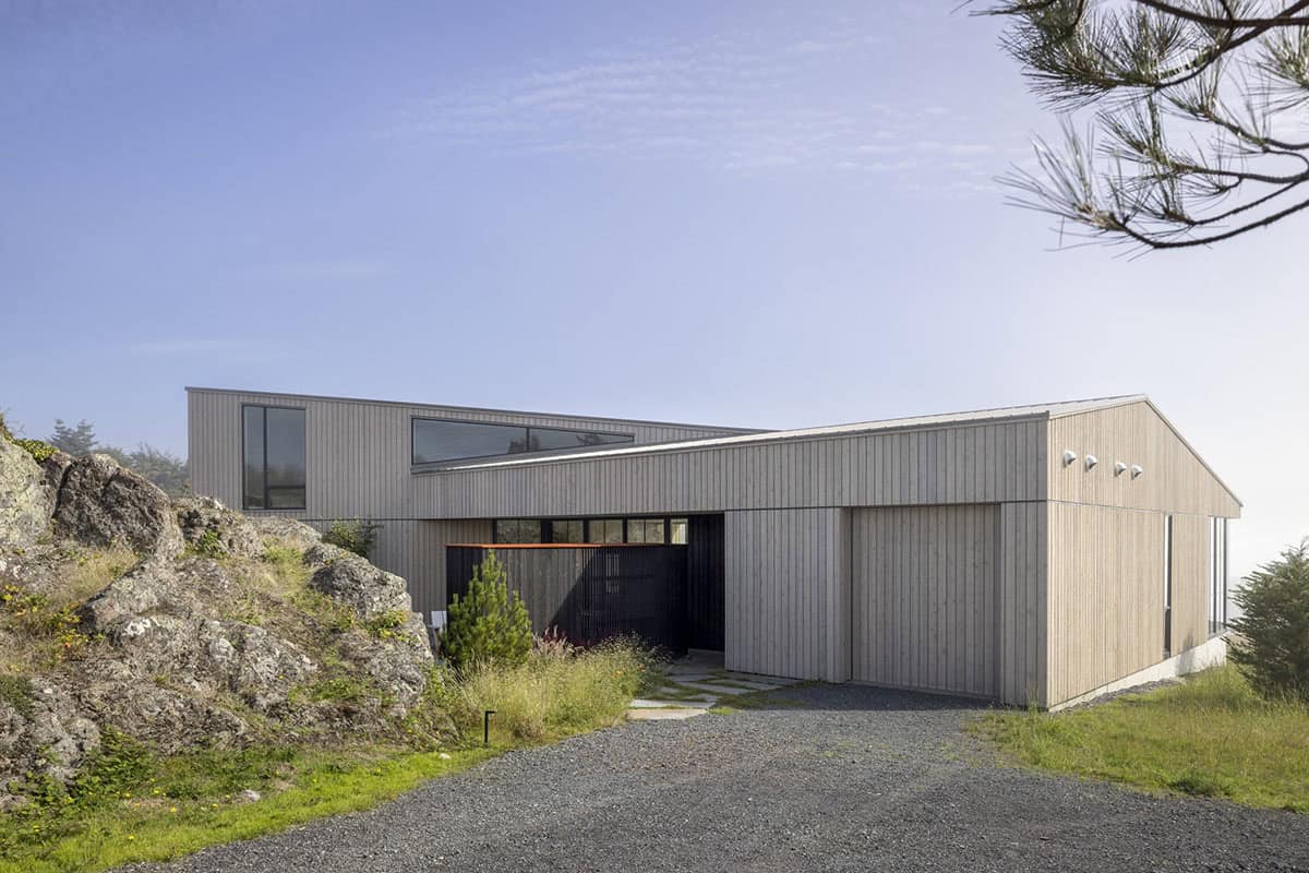 Driveway approach to the long wood-clad house with a garage door and a rock outcrop beside it