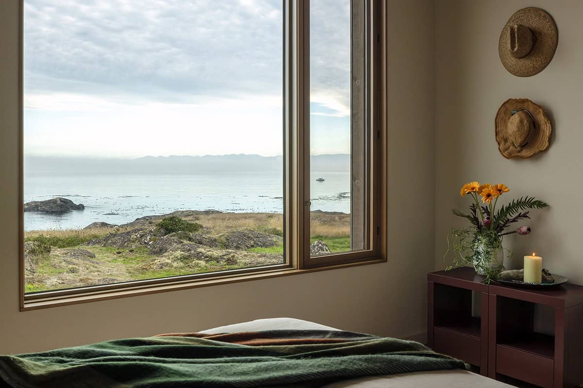 Bedroom window view over rocky shore and water to distant mountains, with hats on the wall and a vase of orange flowers
