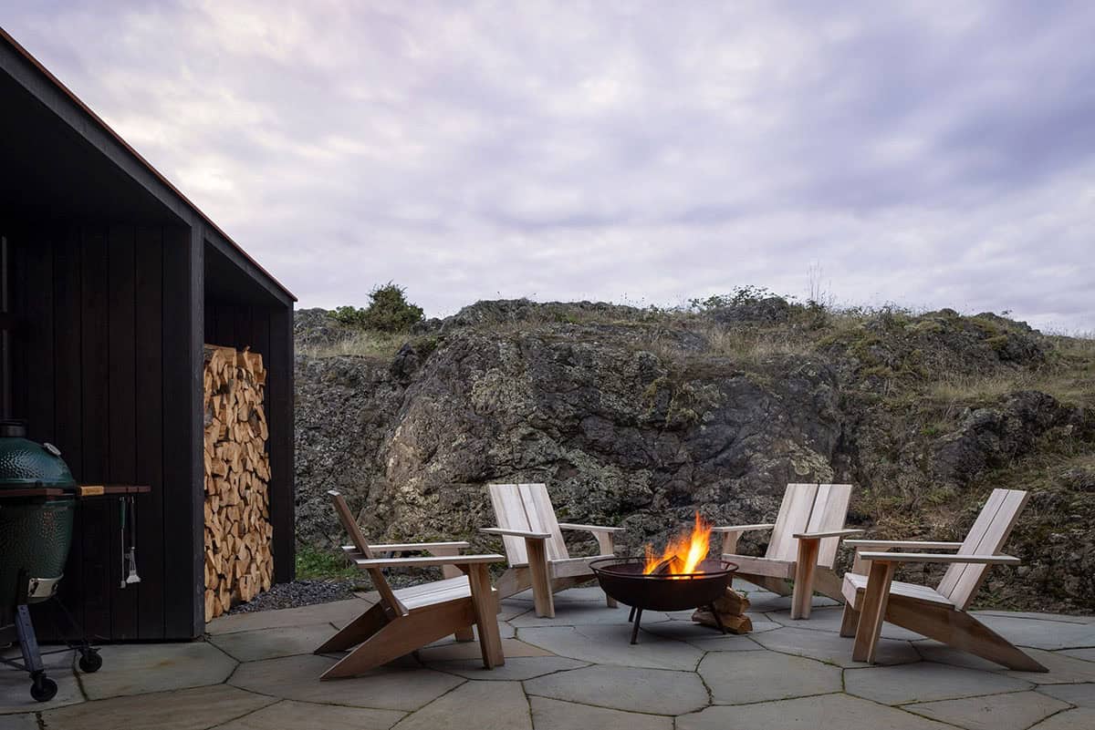 Four Adirondack chairs around a lit fire pit on a flagstone patio, with a firewood shed and a rock outcrop behind