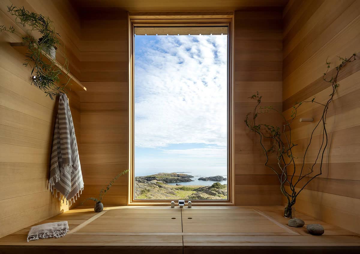 Cedar-lined soaking tub with its wooden cover closed, beneath a tall window framing the rocky shoreline