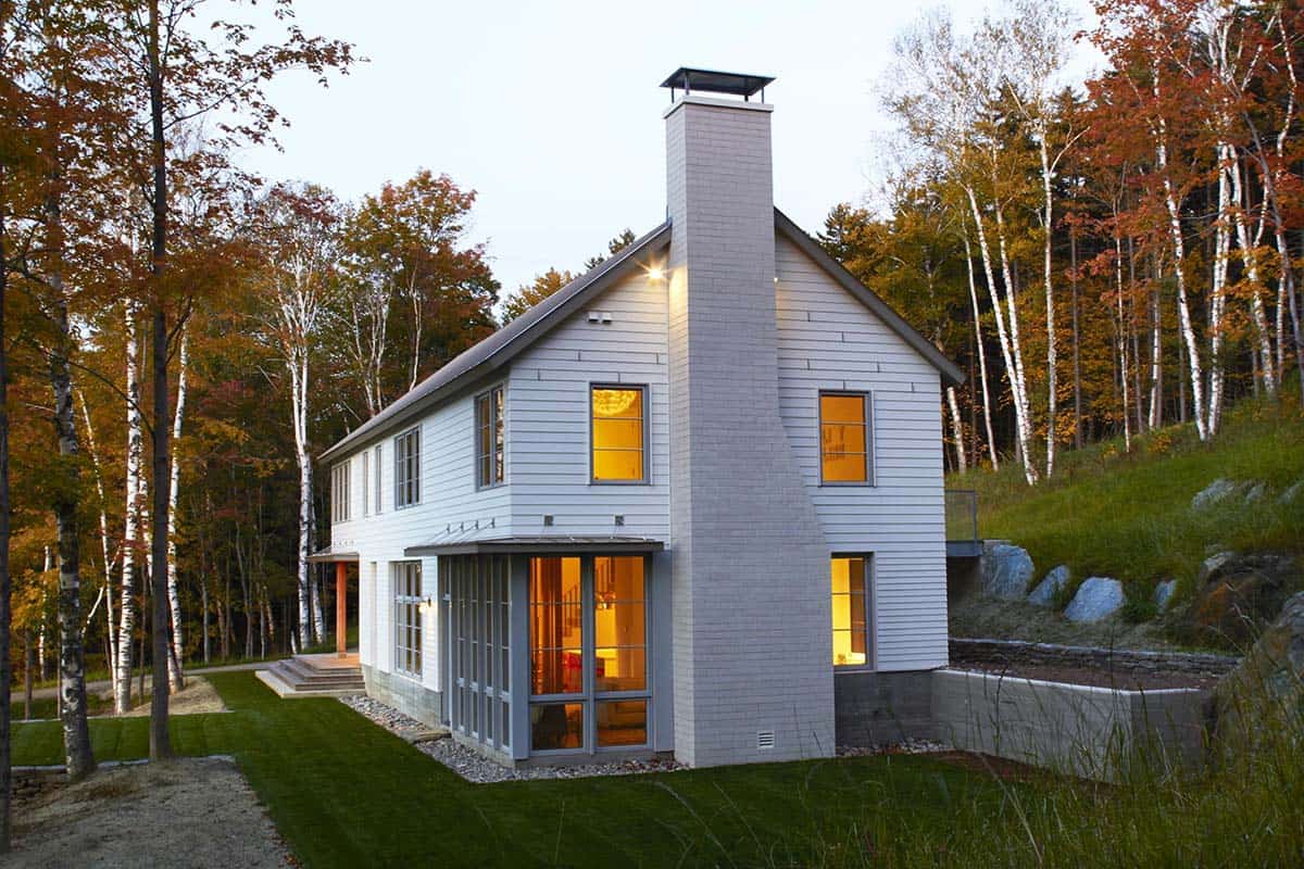 Brick chimney and corner windows lit at dusk among birch trees