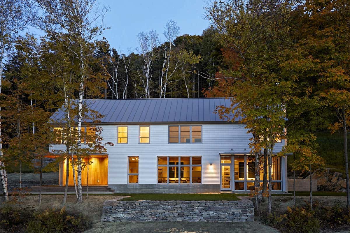 Home at dusk with lit cedar entry and stone wall framed by birch trees