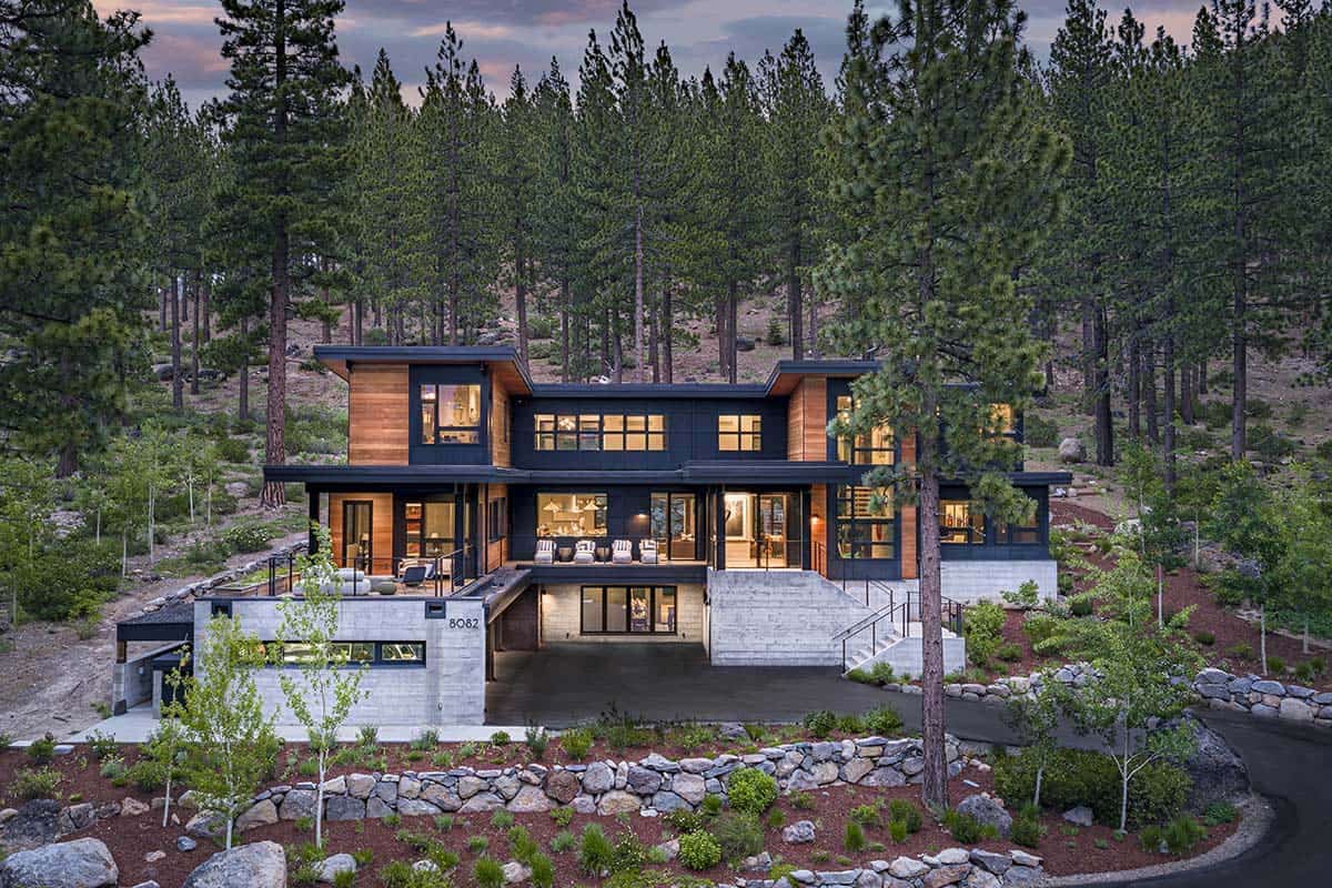 Mountain modern home with cedar and black siding at dusk, glowing windows, and a concrete entry stair among tall pines