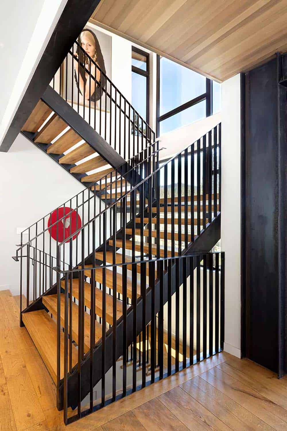 Open-tread wood and black steel staircase with vertical rails, a red circular art piece, and tall windows
