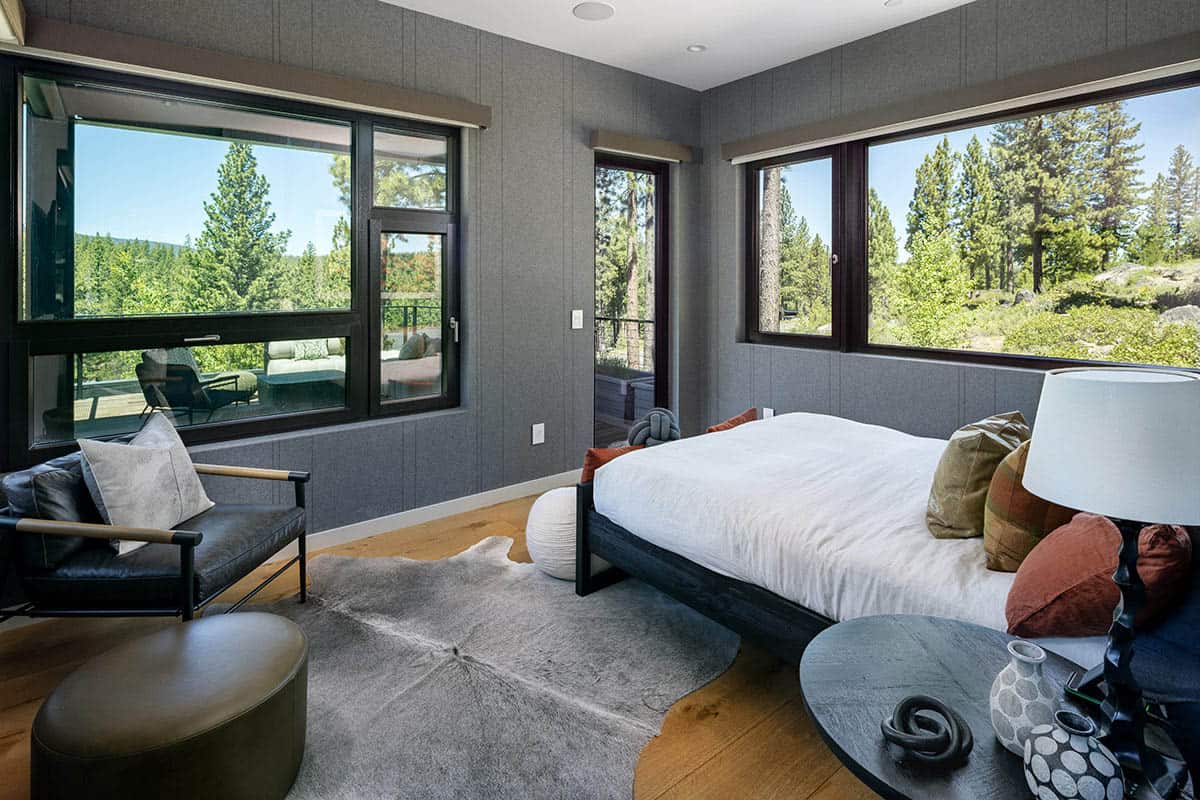 Bedroom with gray fabric-paneled walls, a black bed frame, a cowhide rug, and large forest-view windows