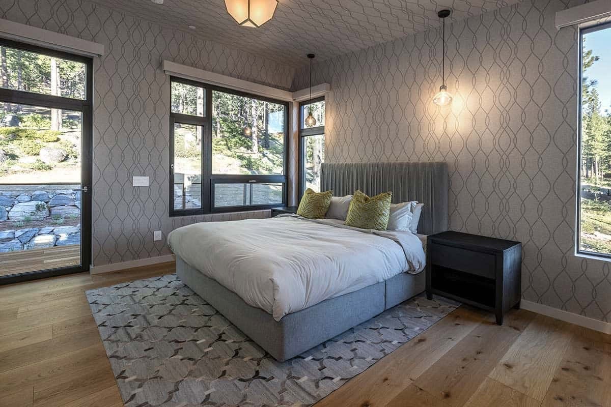 Bedroom with geometric gray wallpaper, an upholstered gray bed, a patterned rug, and forest-view windows