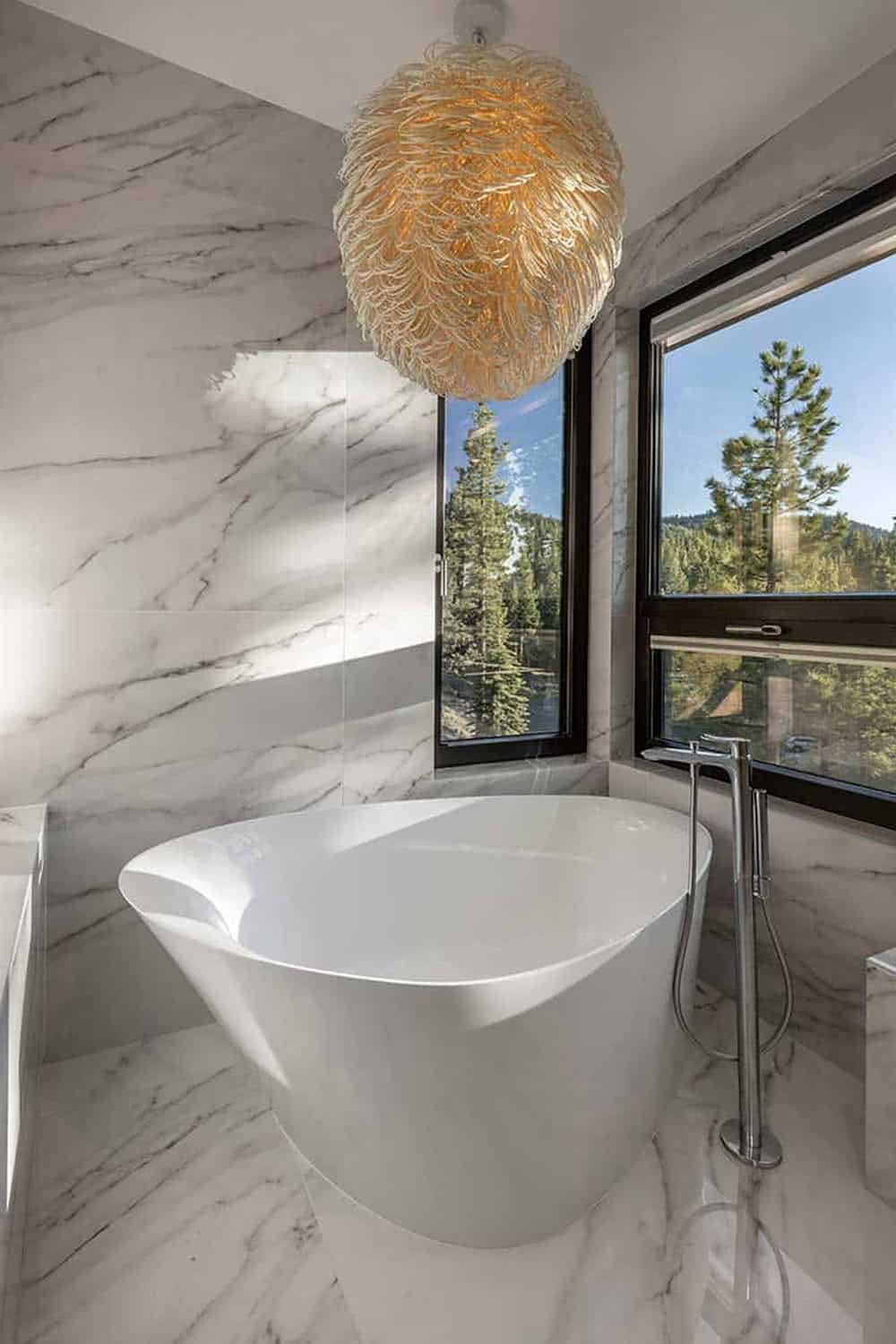 Marble-clad bathroom with a freestanding white tub, a woven pendant light, and corner windows overlooking pines