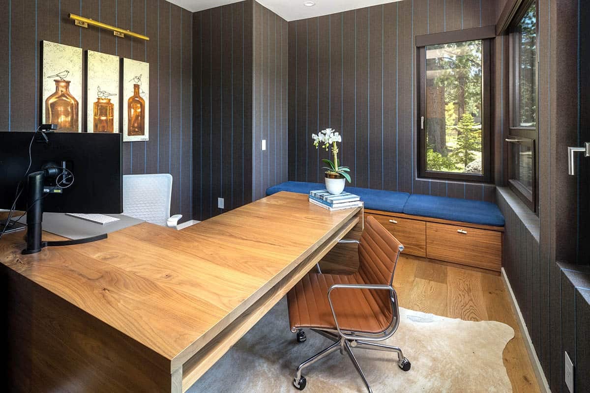 Home office with dark pinstriped walls, a walnut desk, a blue window seat with drawers, and a tan leather chair
