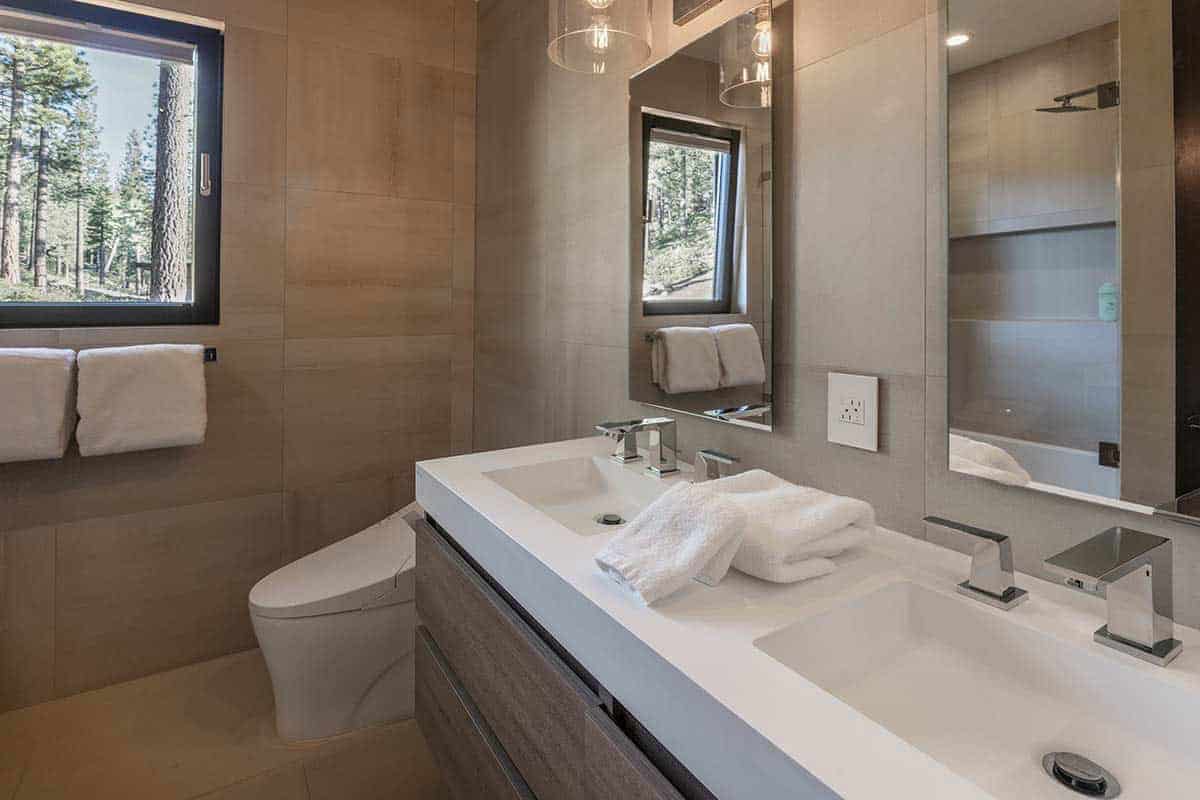 Bathroom with warm beige tile, a white double-sink vanity, a wall-hung toilet, and a window facing the trees