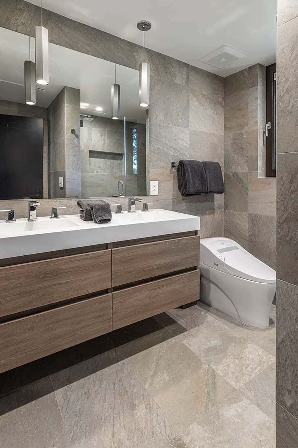 Bathroom with gray stone tile, a floating wood double vanity, linear pendant lights, and a glass-enclosed shower
