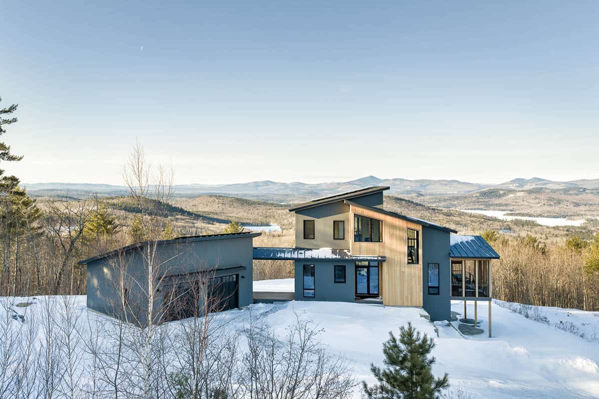 Gray and cedar mountain home with a detached garage on a snowy hilltop, overlooking forested hills and distant peaks