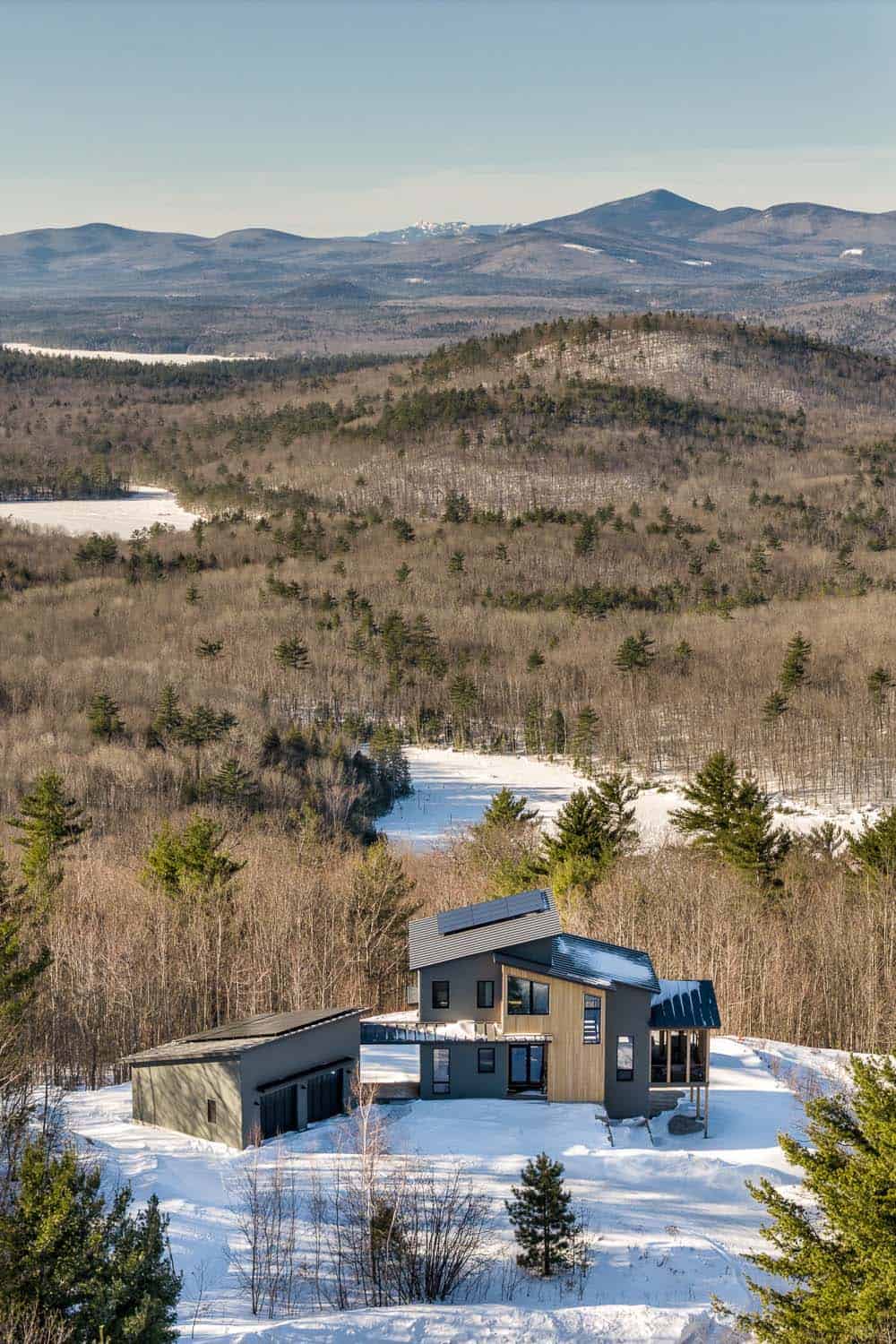 Aerial view of the modern mountain home in snowy woods above a frozen lake and a range of mountains