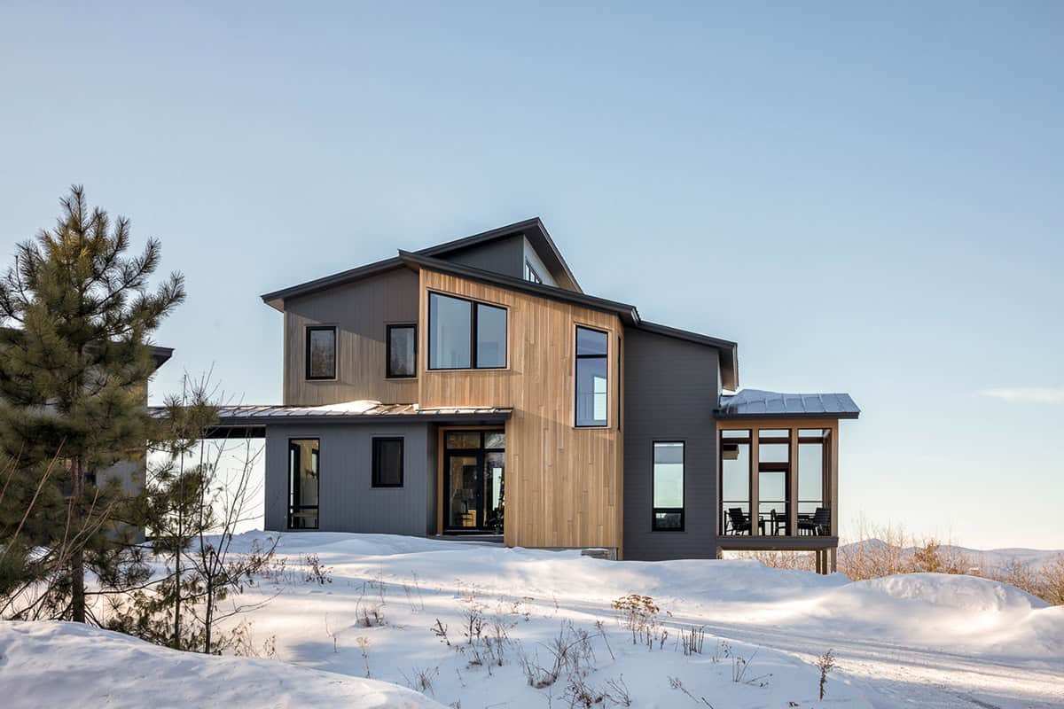 Cedar and charcoal siding with black-framed windows and a screened porch, beside a snow-covered pine