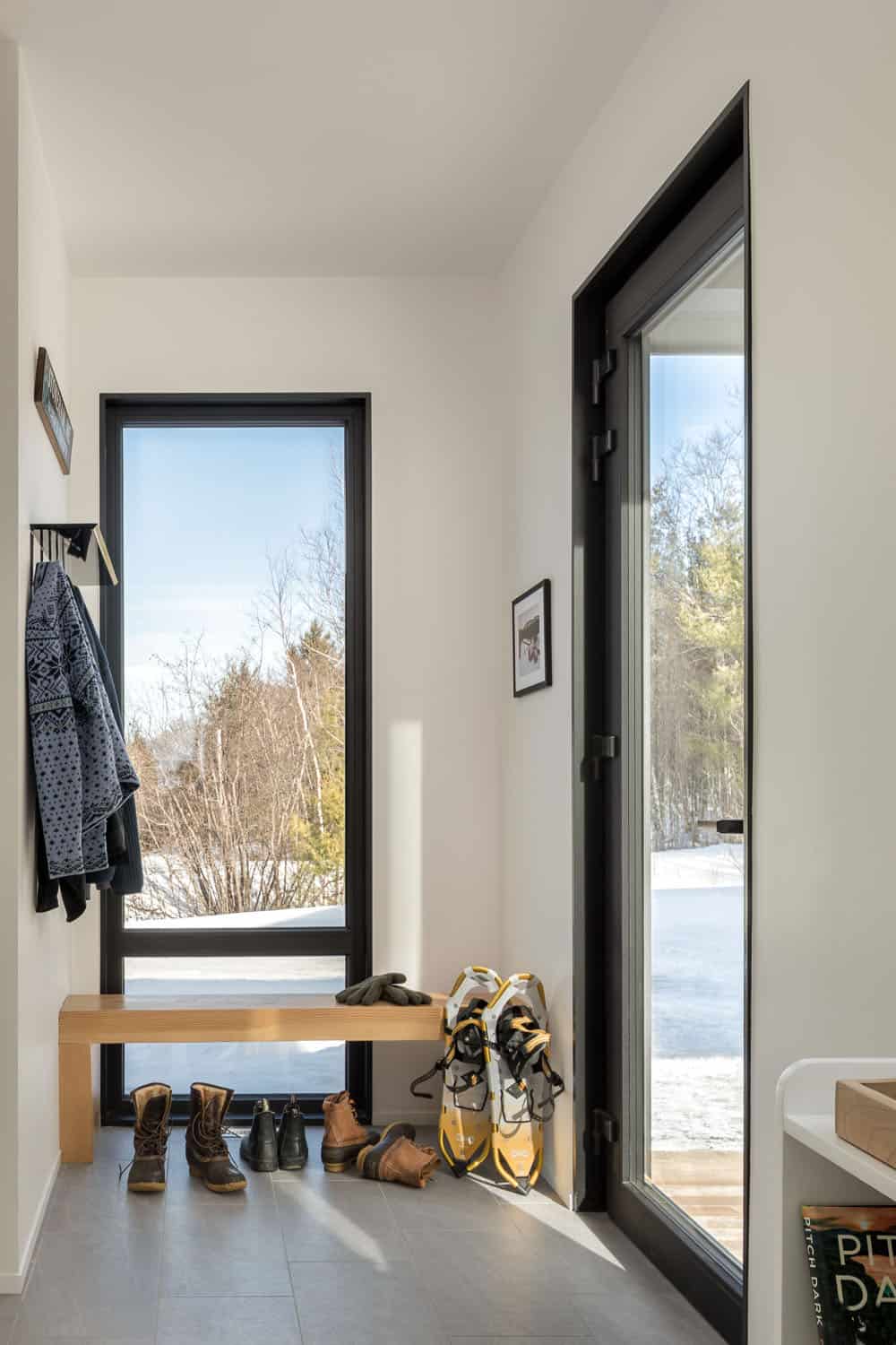 Mudroom with a wood bench, snowshoes, winter boots, and tall black-framed windows onto the snow