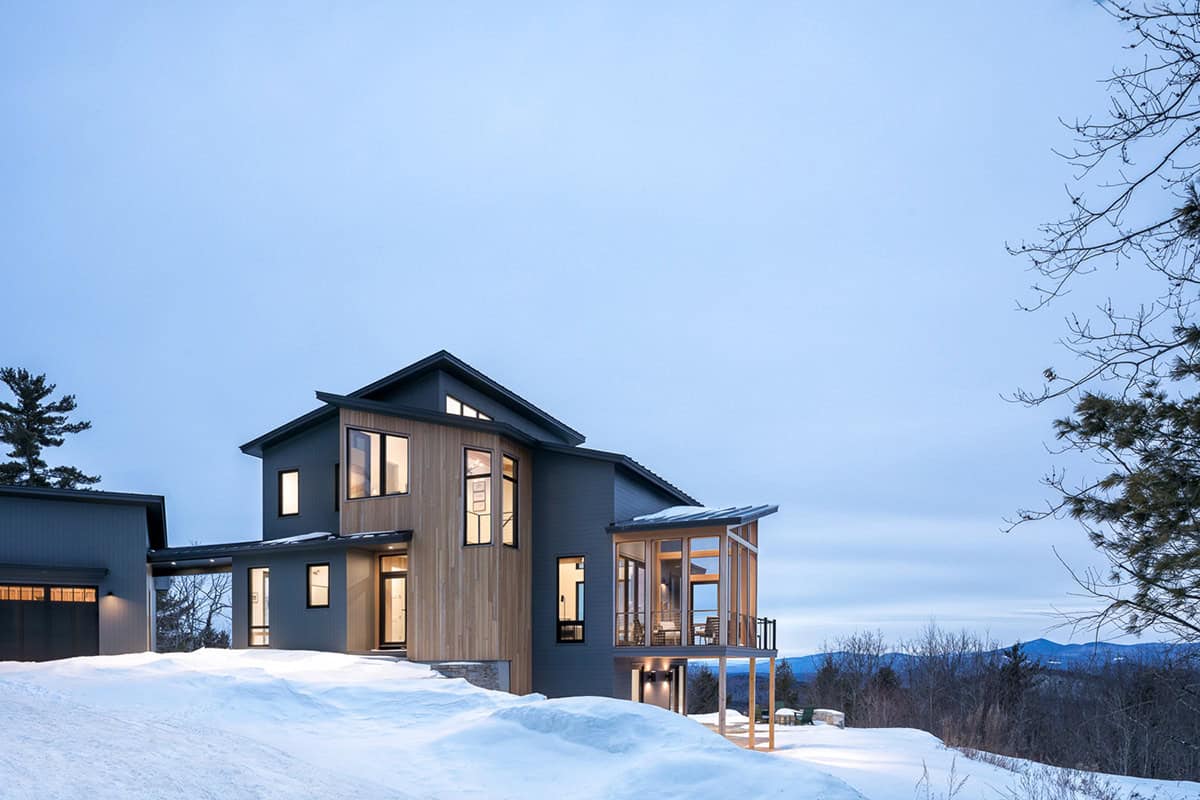 Mountain home at dusk with lit windows, a cedar accent wall, and a screened porch above the snow