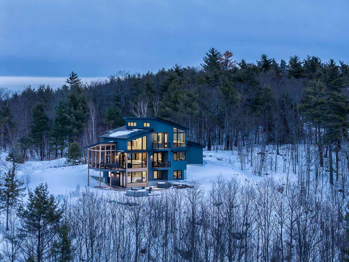 Illuminated three-story home glowing against a dark pine forest at twilight