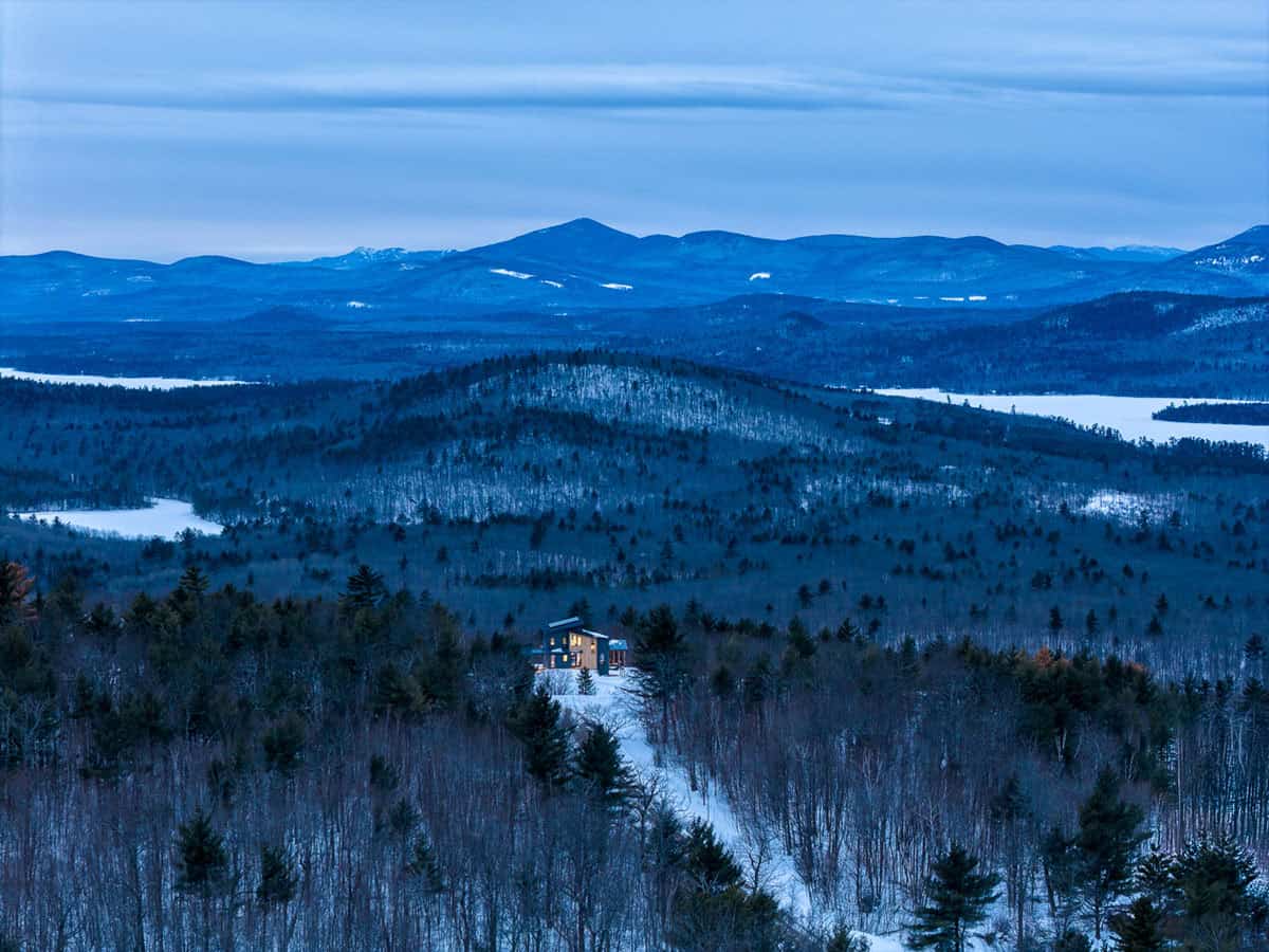Distant glowing home in a snowy forest below layered blue peaks and frozen lakes
