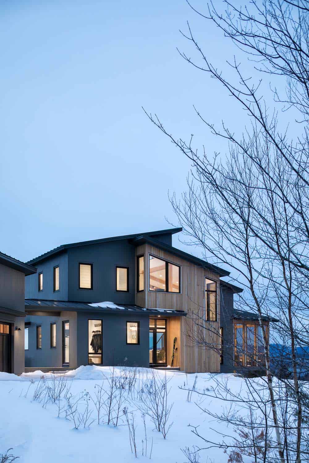 Warmly lit gray and cedar home at dusk with bare birch branches in the foreground