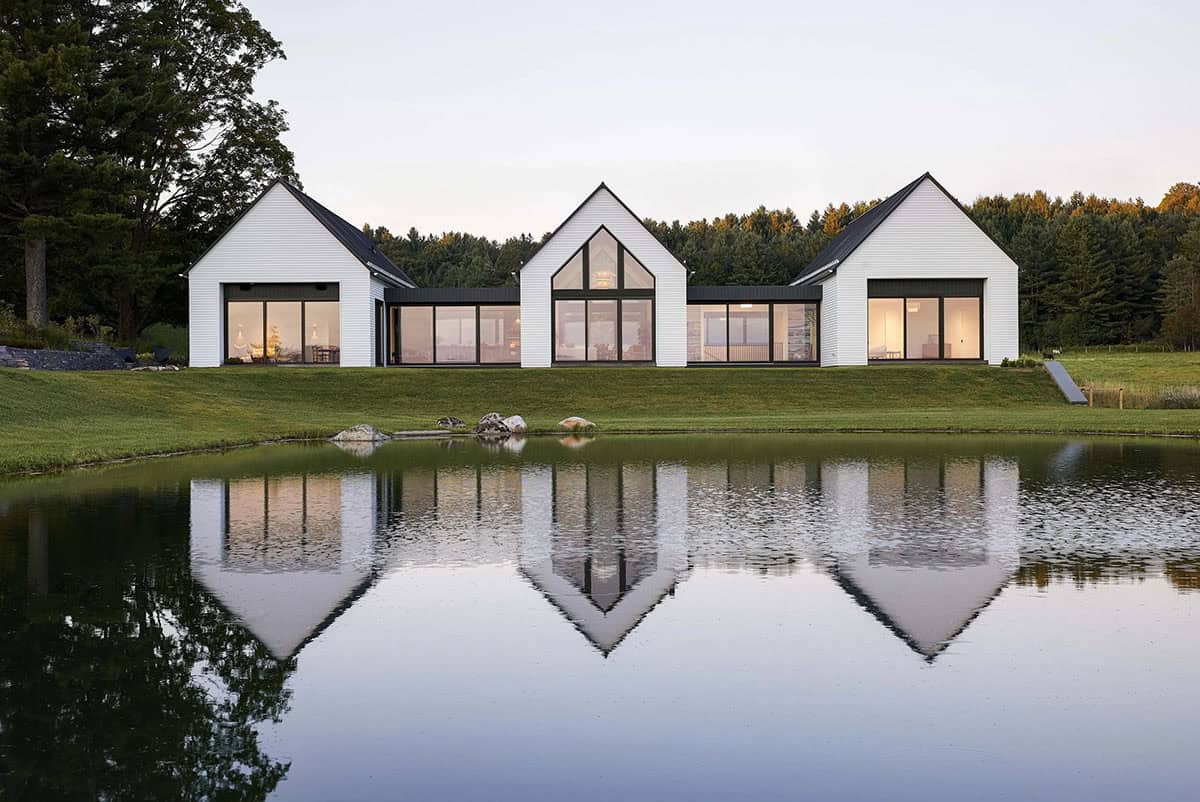 Modern farmhouse in Vermont with three white gables reflected in a pond at dusk