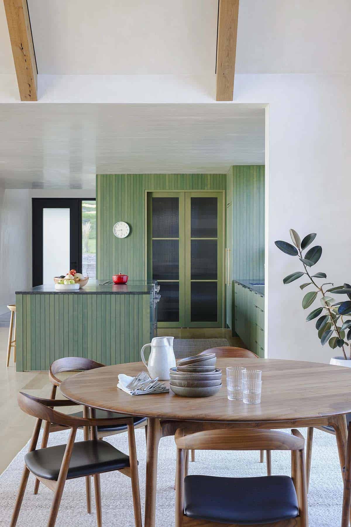 Round walnut dining table with green kitchen and ribbed-glass pantry doors beyond