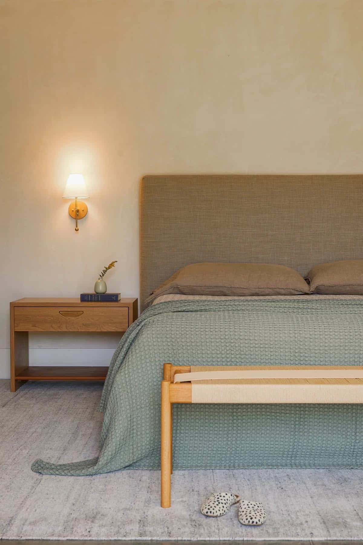 Bedroom detail with sage green bedding, woven bench, and brass sconce