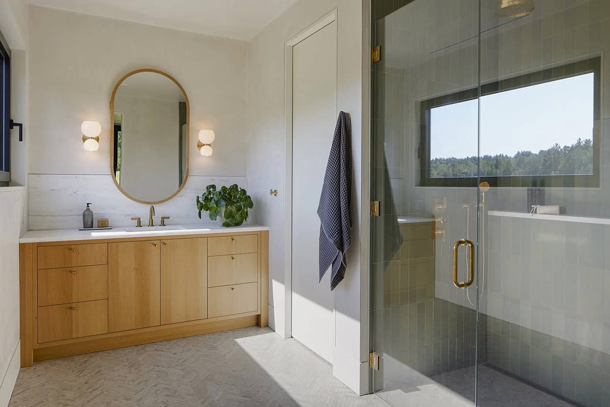 Bathroom with oak vanity, oval mirror, and green tile shower with a window