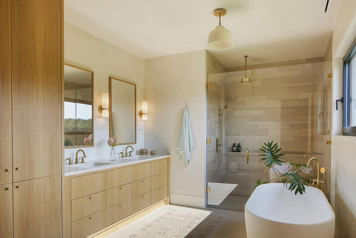 Primary bathroom with oak double vanity, brass fixtures, freestanding tub, and glass shower