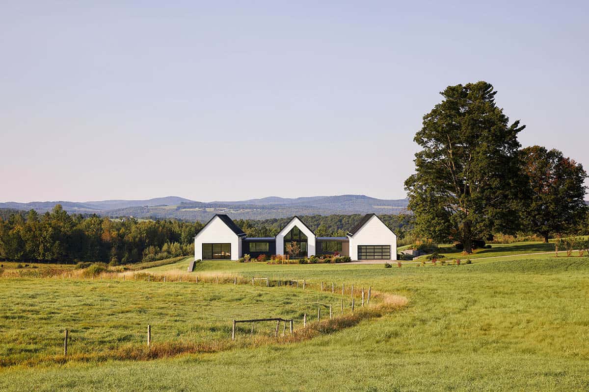 Modern farmhouse with three white gables on a hilltop, with Green Mountains beyond the fields