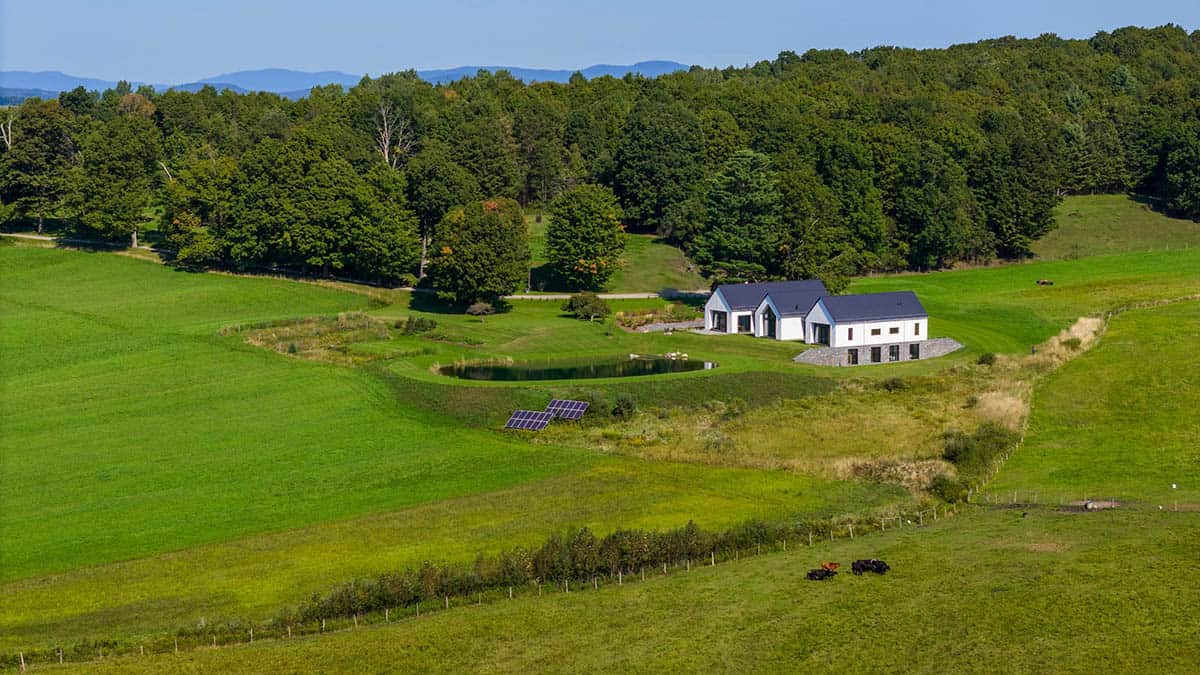 Aerial view of a modern farmhouse by a pond with ground-mounted solar panels in Vermont
