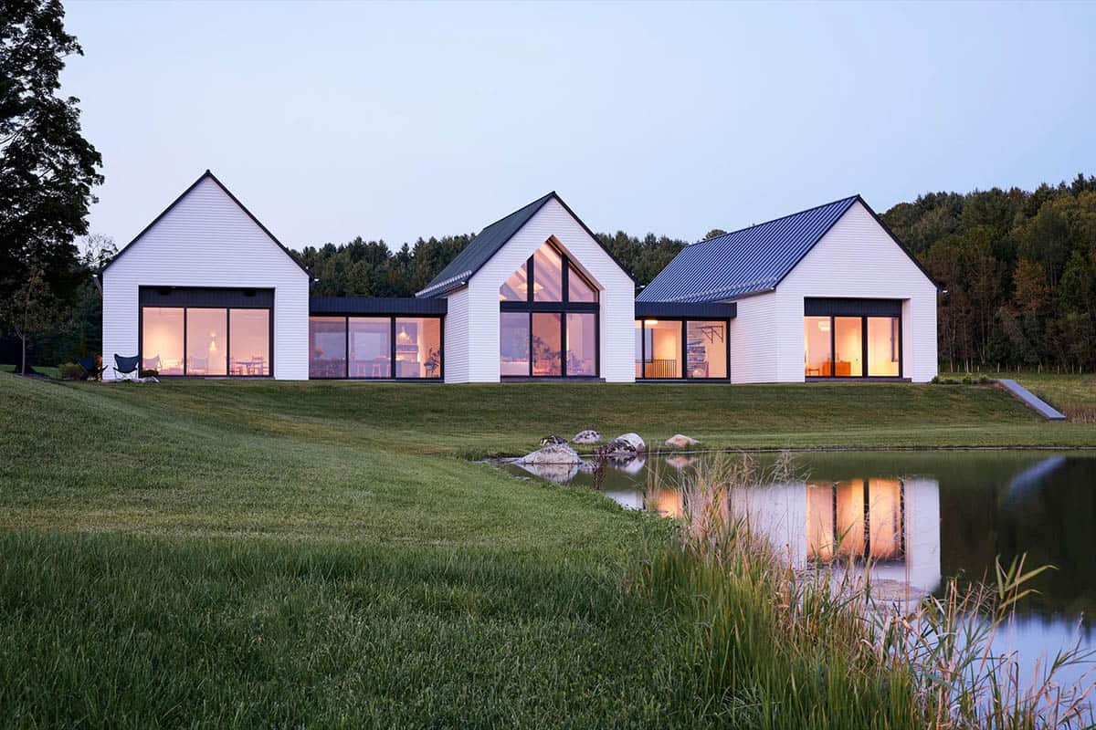 White gabled farmhouse glowing at dusk with pond reflections and large glass windows