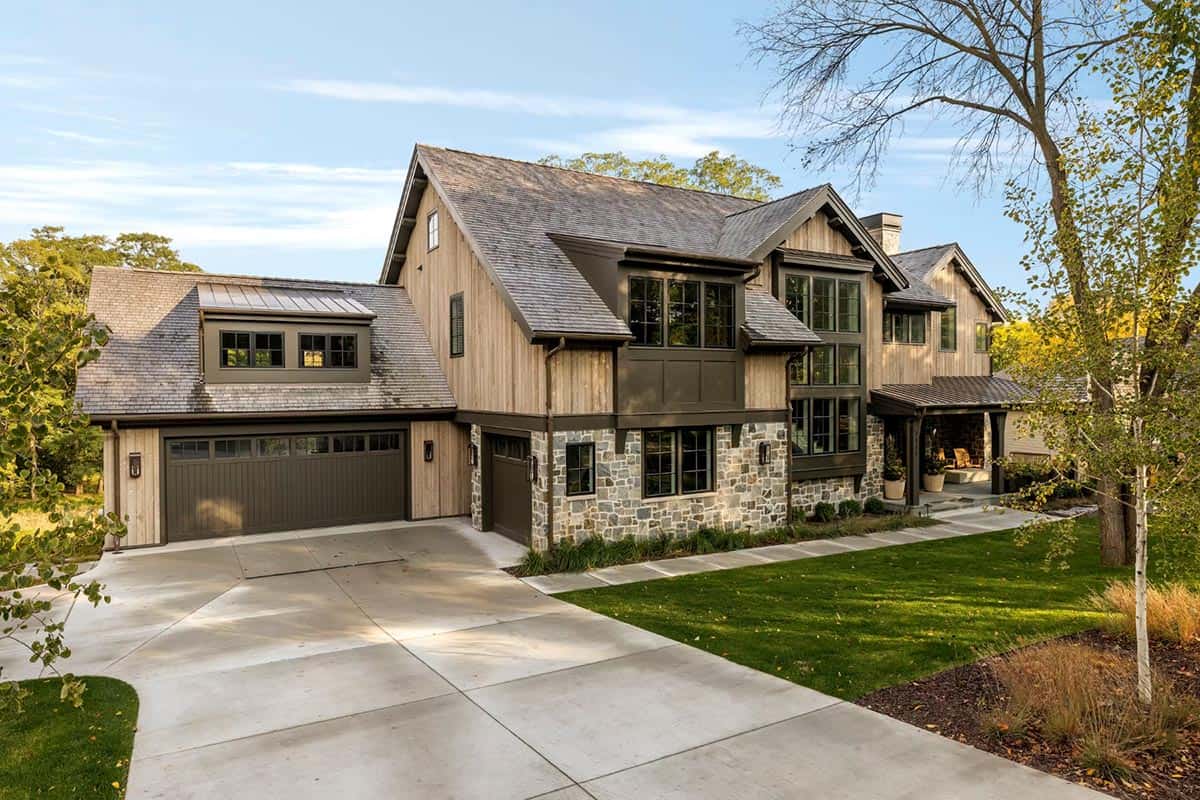 Northwoods-style home with weathered vertical wood siding, a cedar shake roof, natural stone veneer and dark garage doors beside a long concrete driveway