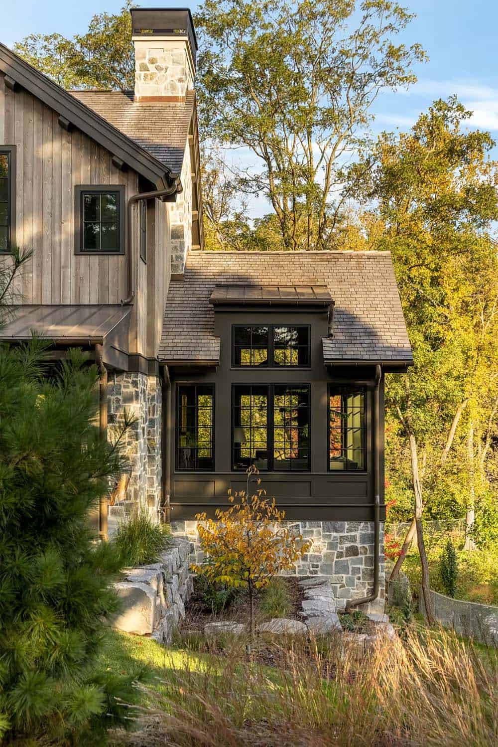 Side of the home with a tall stone chimney, a dark window bay under a metal-capped dormer, and boulder retaining walls planted with native grasses