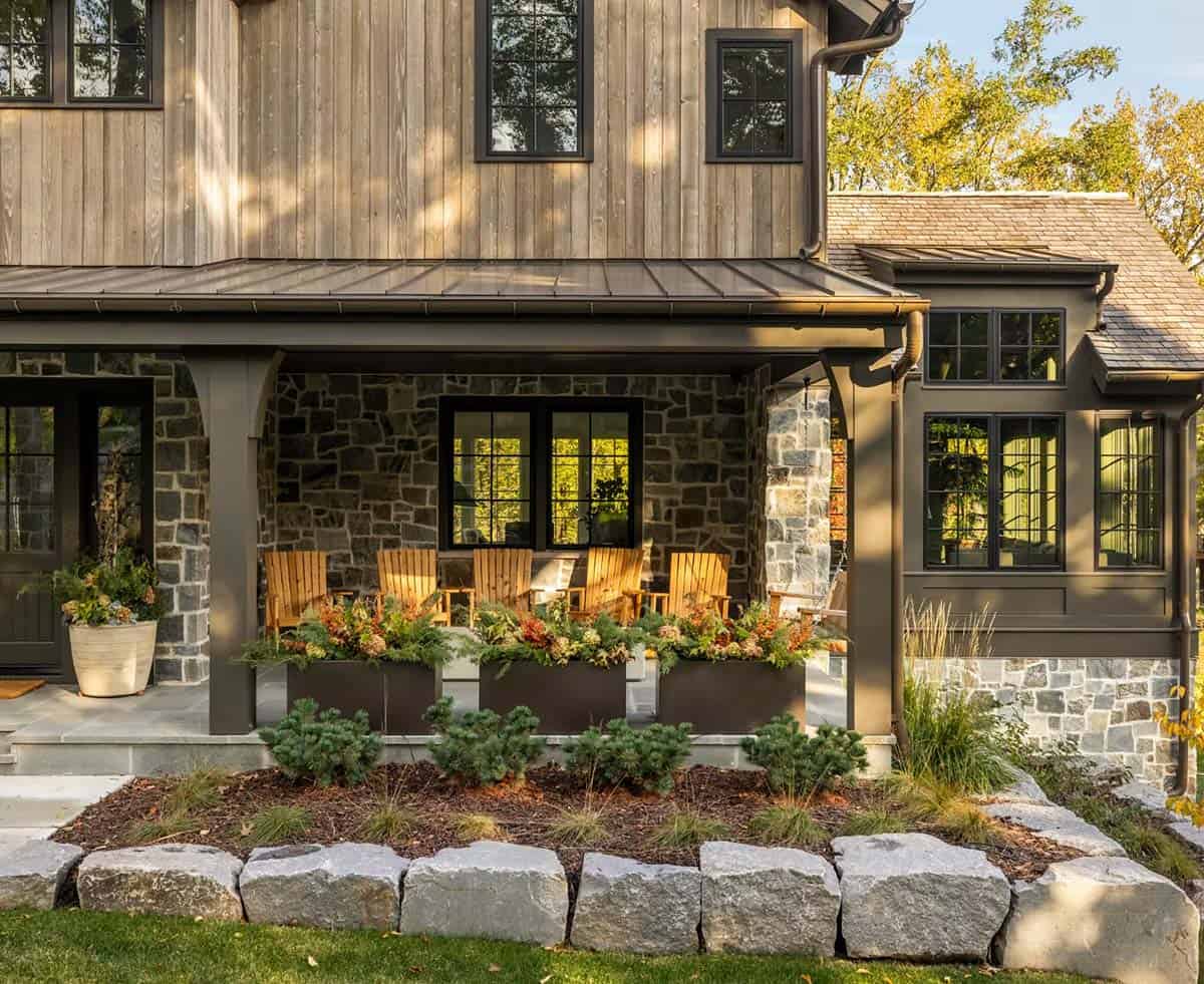 Covered front porch with five wooden Adirondack chairs, black planters of fall flowers and a stone wall, edged by granite boulders and dwarf pines