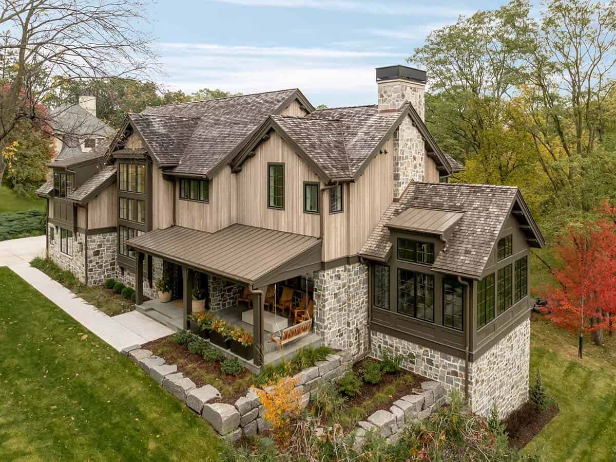 Aerial view of the home's layered gables, metal porch roof, stone chimney and window-wrapped wing surrounded by fall trees