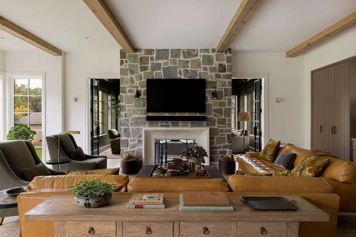 Living room with a stone fireplace wall and limestone surround, a cognac leather sectional, olive velvet armchairs and rustic wood ceiling beams