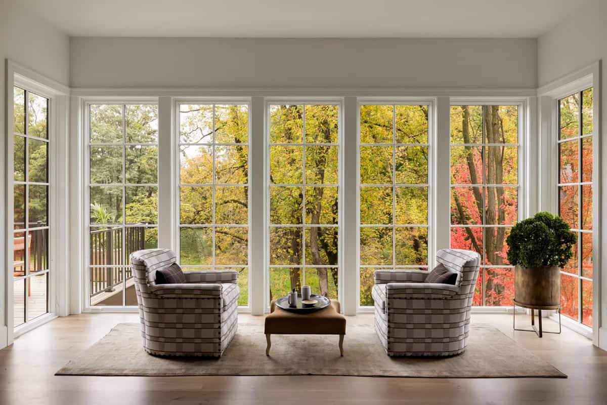 Sitting area with two plaid armchairs and a leather ottoman in front of a wall of tall grid windows framing yellow and red fall trees