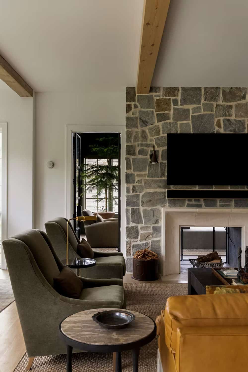 Olive velvet armchairs and a brass floor lamp beside a stone fireplace wall, with a doorway view into a green-paneled family room